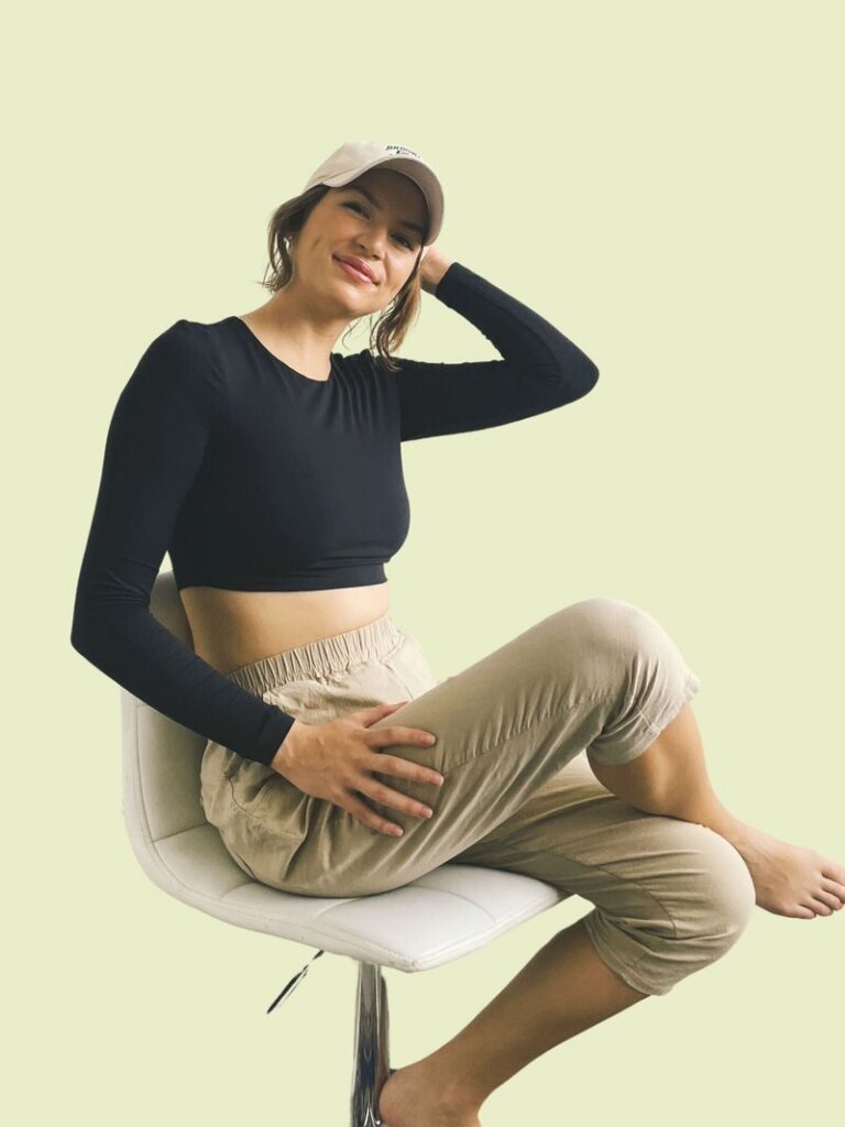 Caitlin Christensen posing on a chair in brown pant baseball cap and black top. 